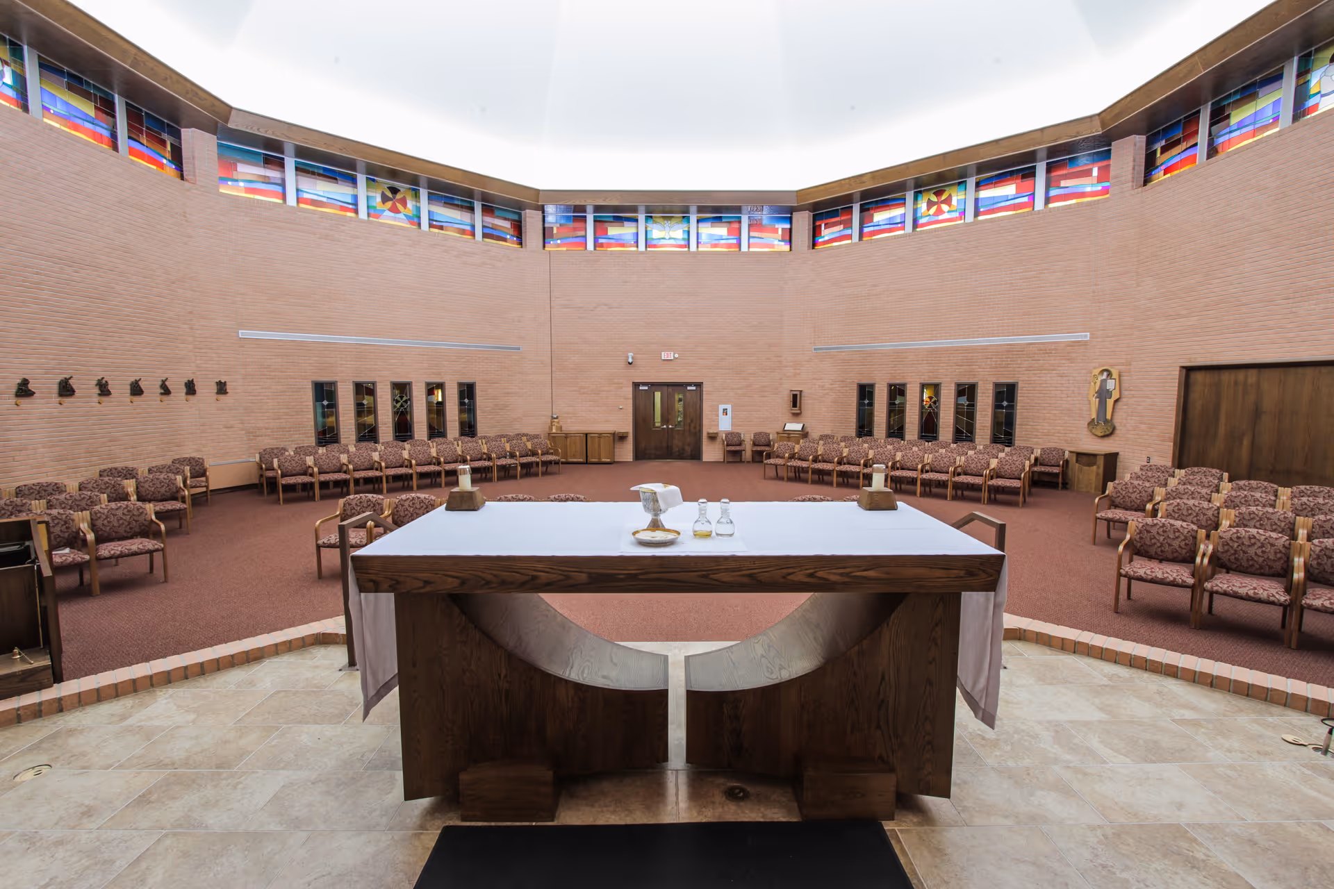 Interior view of a chapel or worship space with a wooden altar covered with a white cloth in the foreground. The room has a circular arrangement of patterned chairs along the walls, brick walls, and stained glass windows near the ceiling.