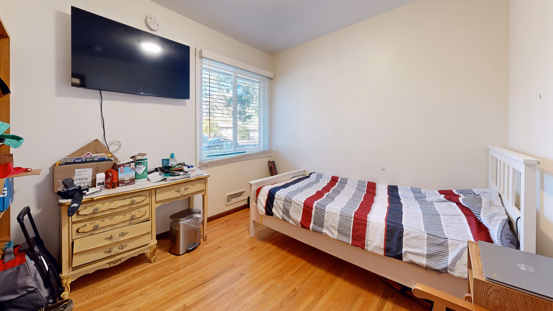 A small bedroom with a single bed covered in a striped red, white, gray, and black comforter. There is a wooden dresser with various items on top, including boxes and bottles, beneath a wall-mounted flat-screen TV. A window with blinds lets in natural light, and the floor is wooden. A laptop is placed on a small table next to the bed.