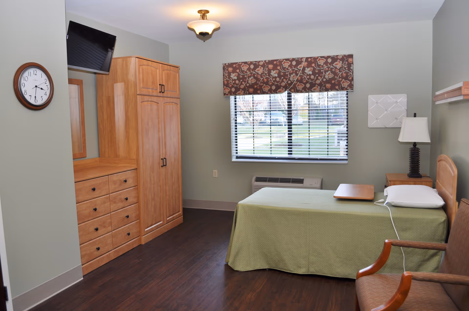 A simple bedroom with a single bed covered with a green bedspread, a wooden headboard, and a pillow. There is a wooden nightstand with a lamp next to the bed, a wooden dresser with multiple drawers, and a tall wooden wardrobe. A wall-mounted TV is positioned above the dresser. A window with blinds and a floral valance lets in natural light. The room has light green walls and dark wood flooring.