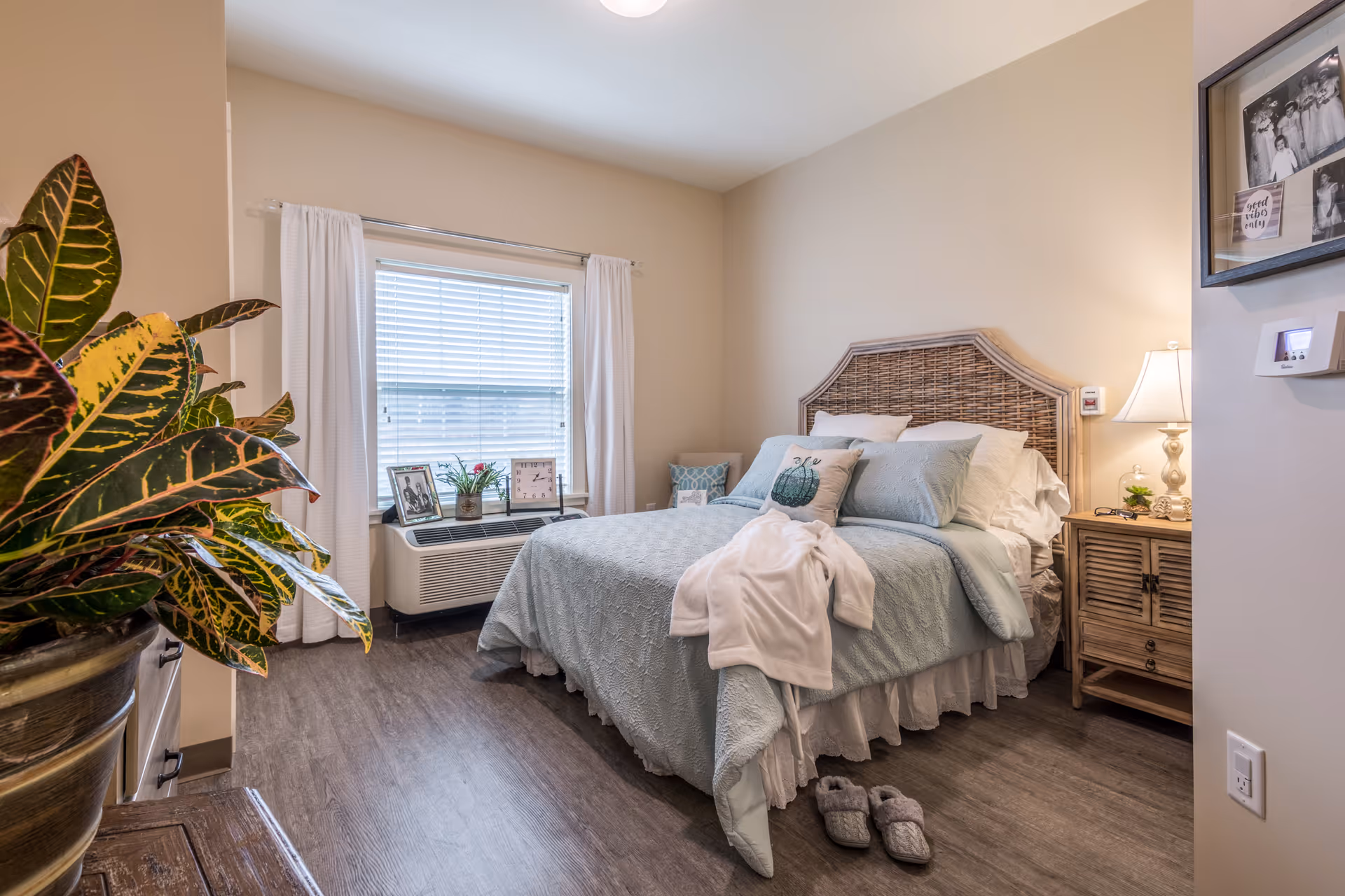 A cozy bedroom with a neatly made bed featuring light blue bedding and decorative pillows. A white robe is draped over the bed, and a pair of slippers are placed on the floor nearby. There is a wooden nightstand with a lamp and decorative items on the right side of the bed. A window with white curtains and blinds is behind the bed, and a large leafy plant is visible on the left side of the image. The room has wood flooring and neutral-colored walls.