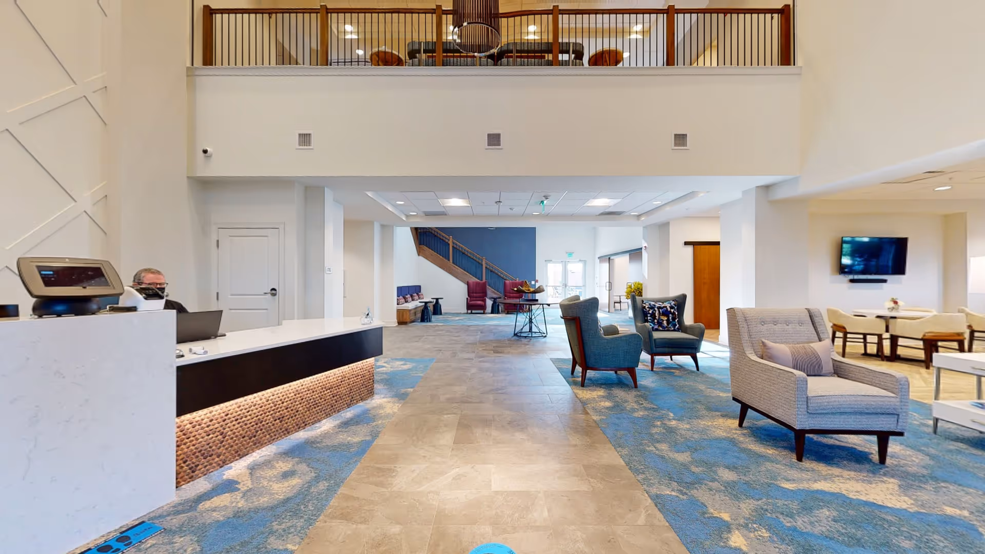 Spacious and modern assisted living facility lobby with a reception desk on the left, a person sitting behind the desk, comfortable seating areas with armchairs and sofas on the right, a staircase at the back, and a second-floor balcony overlooking the lobby.