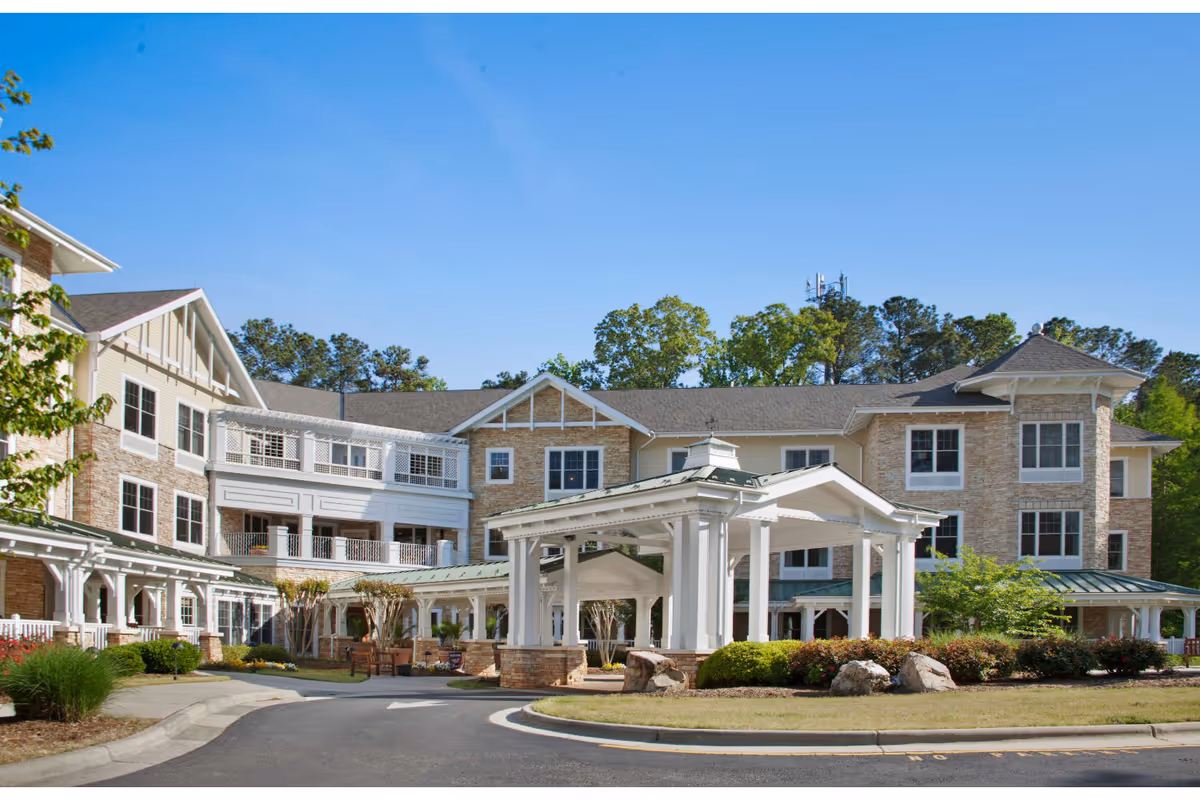 Exterior view of a multi-story senior living facility building with a covered entrance, surrounded by landscaped greenery and trees under a clear blue sky.