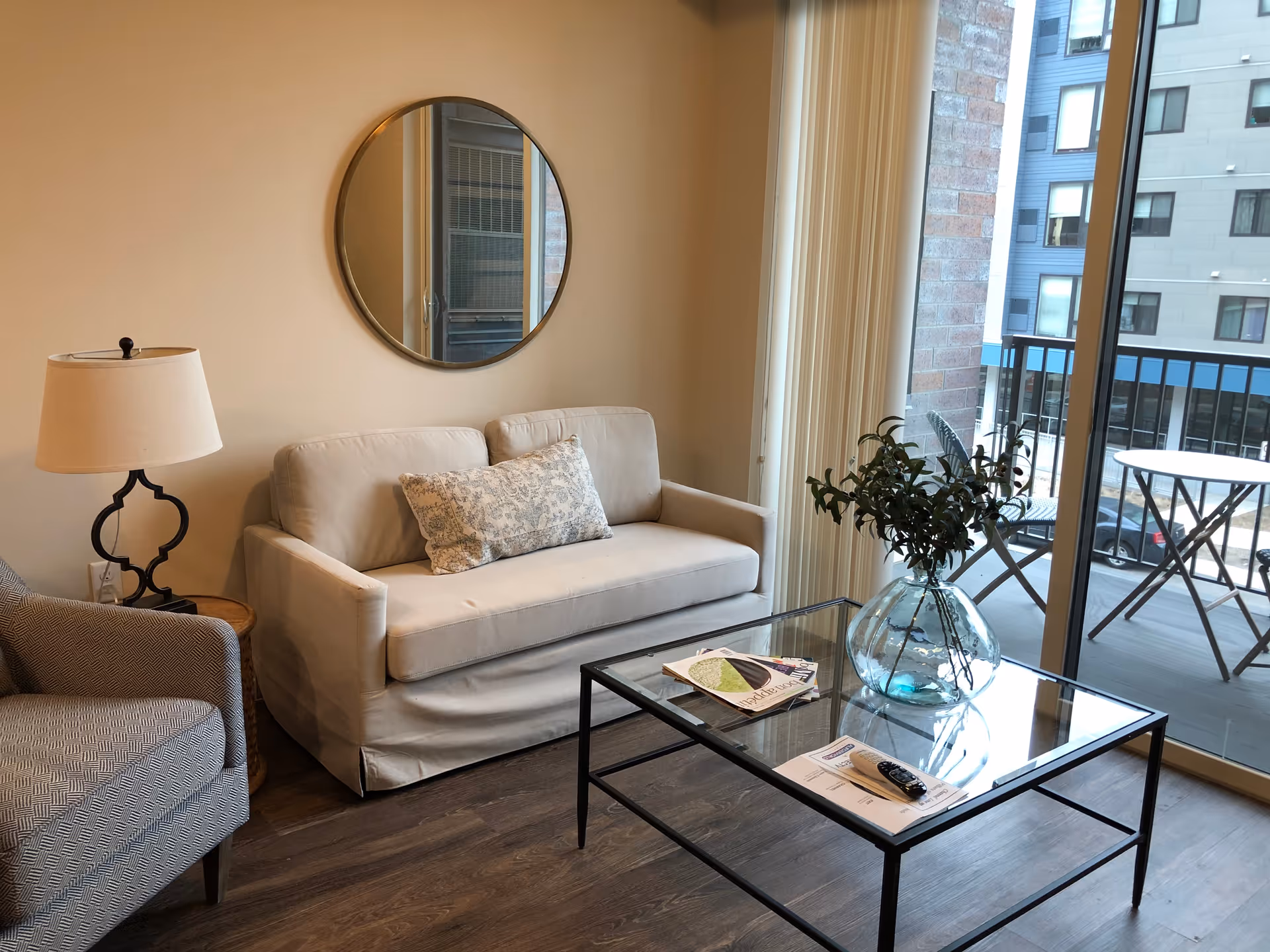 Cozy living room featuring a beige loveseat, armchair, glass coffee table with a vase, and sliding glass doors opening to a small balcony.