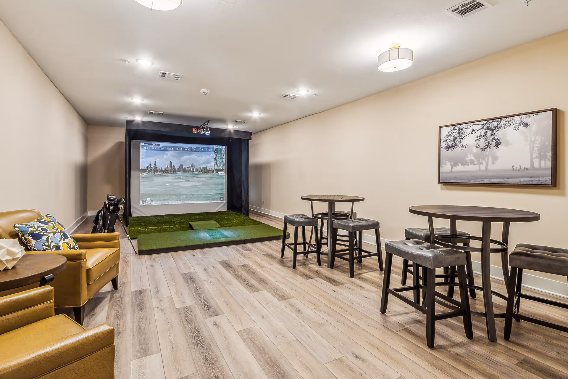 A room with a golf simulator setup at the far end, featuring a large screen displaying a golf course. The room has light wood flooring, beige walls, two round tables with four cushioned stools, and two tan leather armchairs with a small side table. A framed black and white picture of people playing golf hangs on the wall.