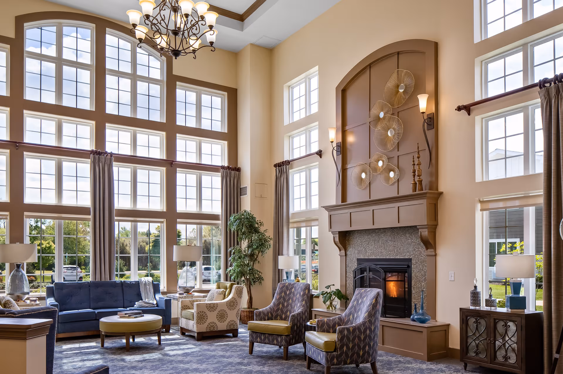 A spacious and well-lit living room with large floor-to-ceiling windows allowing natural light to fill the space. The room features a blue sofa, patterned armchairs, a round ottoman, and a fireplace with decorative wall art above it. There are also several lamps and a potted plant, creating a cozy and inviting atmosphere.