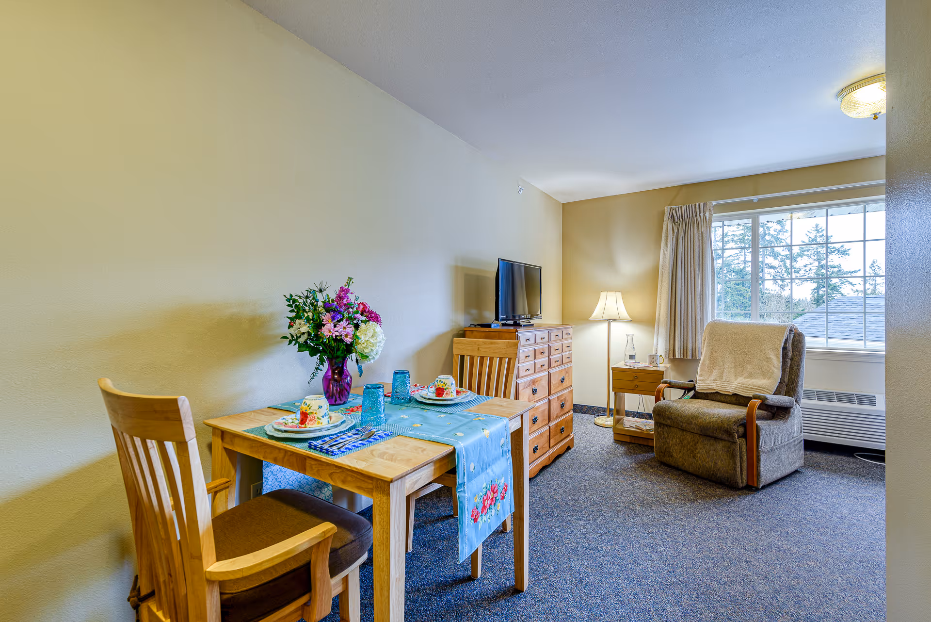 Cozy assisted living room featuring a set dining table, wooden dresser with a TV, and a recliner by a window.