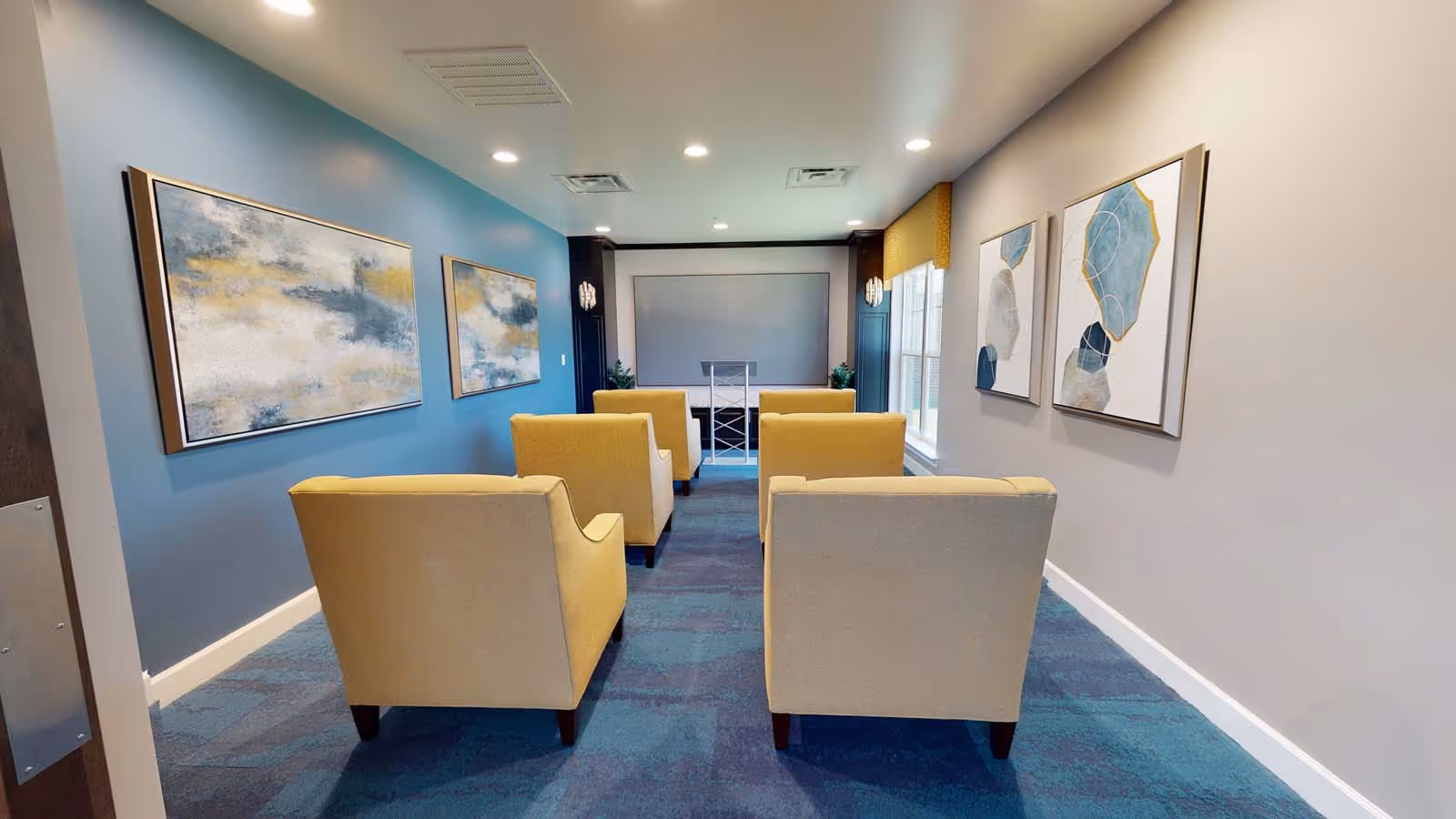 A small conference or media room with four beige armchairs arranged in two rows facing a screen at the front. The room has blue carpet, blue and gray walls adorned with abstract paintings, and windows on the right side letting in natural light.