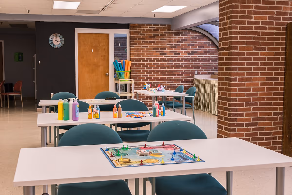 A senior living facility activity room with white tables and teal chairs. On the tables are various craft supplies including bottles of paint, glue, and a board game. The room has brick walls, a wooden door, a clock on a dark wall, and a rack holding colorful pool noodles.