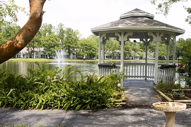 A peaceful outdoor scene featuring a white wooden gazebo on a wooden deck overlooking a pond with a water fountain. Surrounding the pond are green trees and shrubs, with a paved walkway and a birdbath in the foreground.