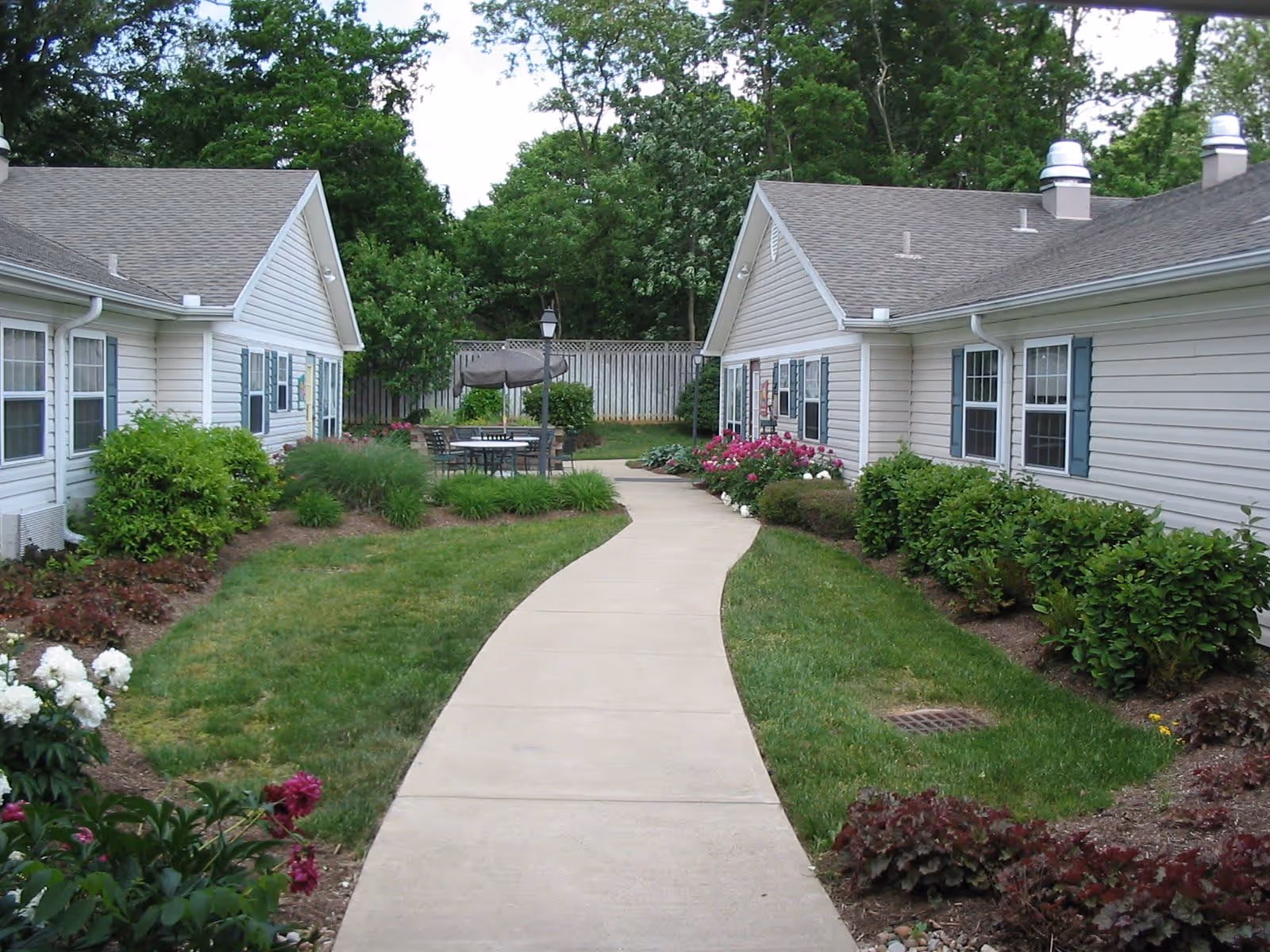 A paved walkway flanked by well-maintained green lawns and flower beds leads between two single-story buildings with beige siding and blue shutters. At the end of the path, there is an outdoor seating area with a table, chairs, and a closed umbrella, surrounded by trees and a wooden fence.