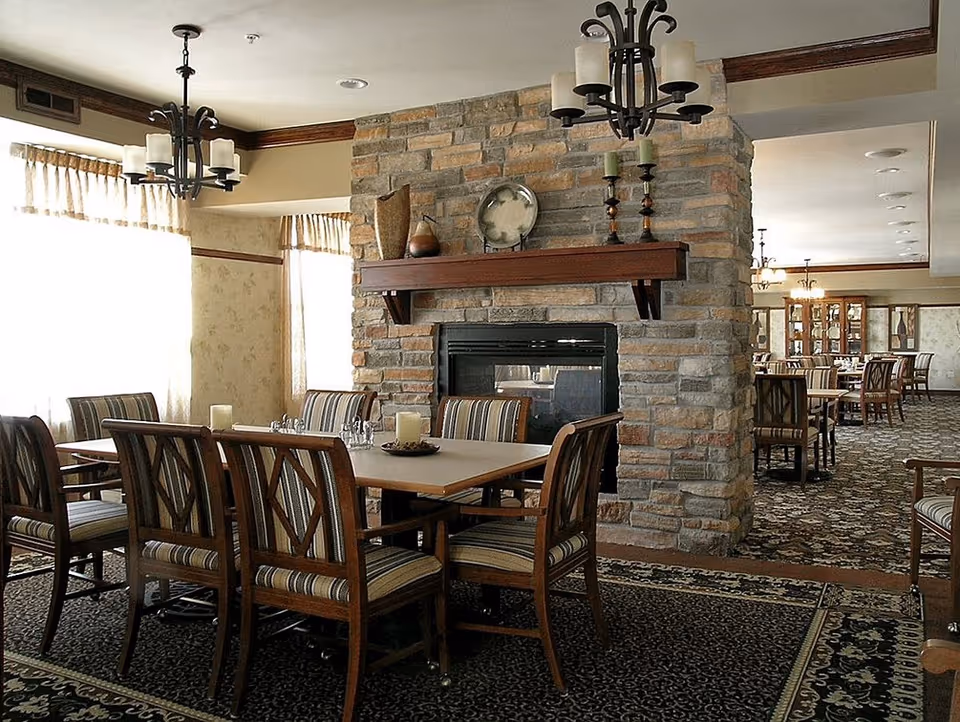 A dining area in a senior living facility with wooden tables and chairs featuring striped cushions. The room has a stone fireplace with decorative items on the mantel, including candles and a plate. Large windows with curtains allow natural light to fill the space. The carpet has a floral pattern, and chandeliers hang from the ceiling.