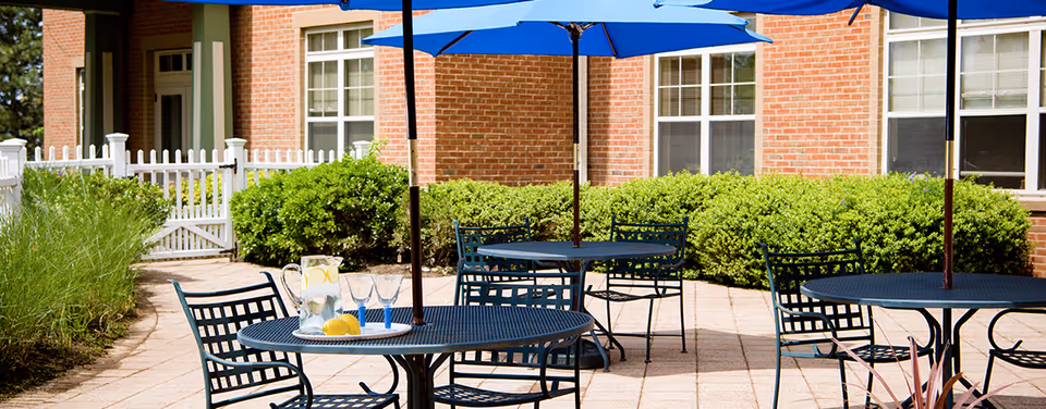 Outdoor patio with metal tables and blue umbrellas beside a brick building and landscaping.