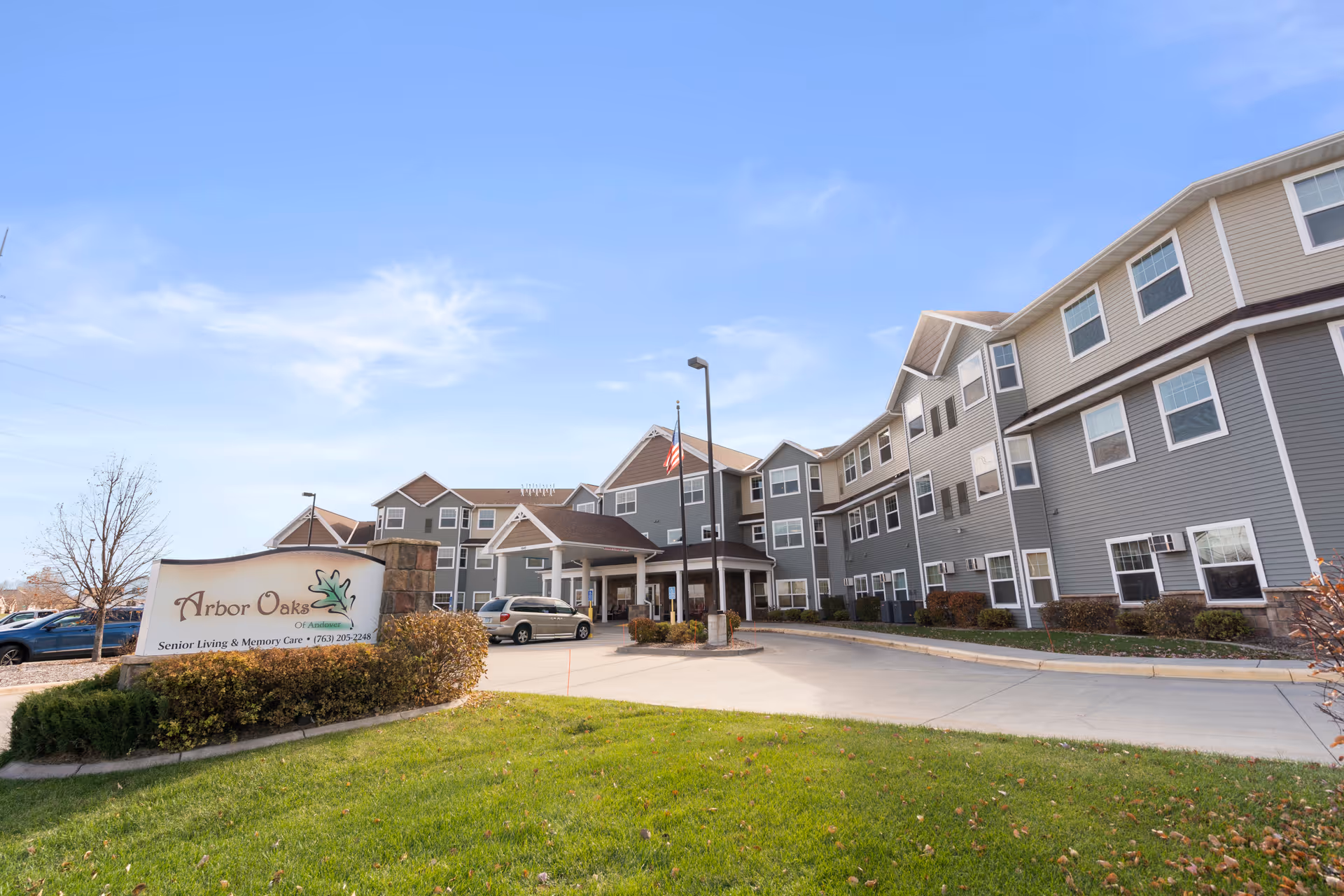Front exterior of a multi-story senior living building with a driveway, entrance canopy, American flag, and an 'Arbor Oaks' sign on the lawn.