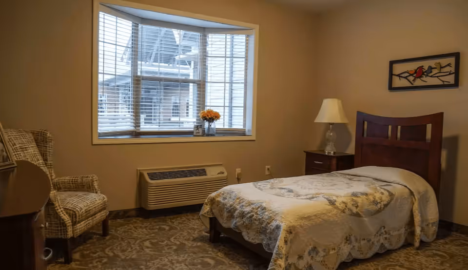 A cozy bedroom with a single bed covered in a patterned quilt, a wooden headboard, a nightstand with a lamp, a patterned armchair, and a window with blinds letting in natural light. A small vase with flowers and a framed photo are on the windowsill, and a decorative wall art featuring birds on a branch hangs above the bed.