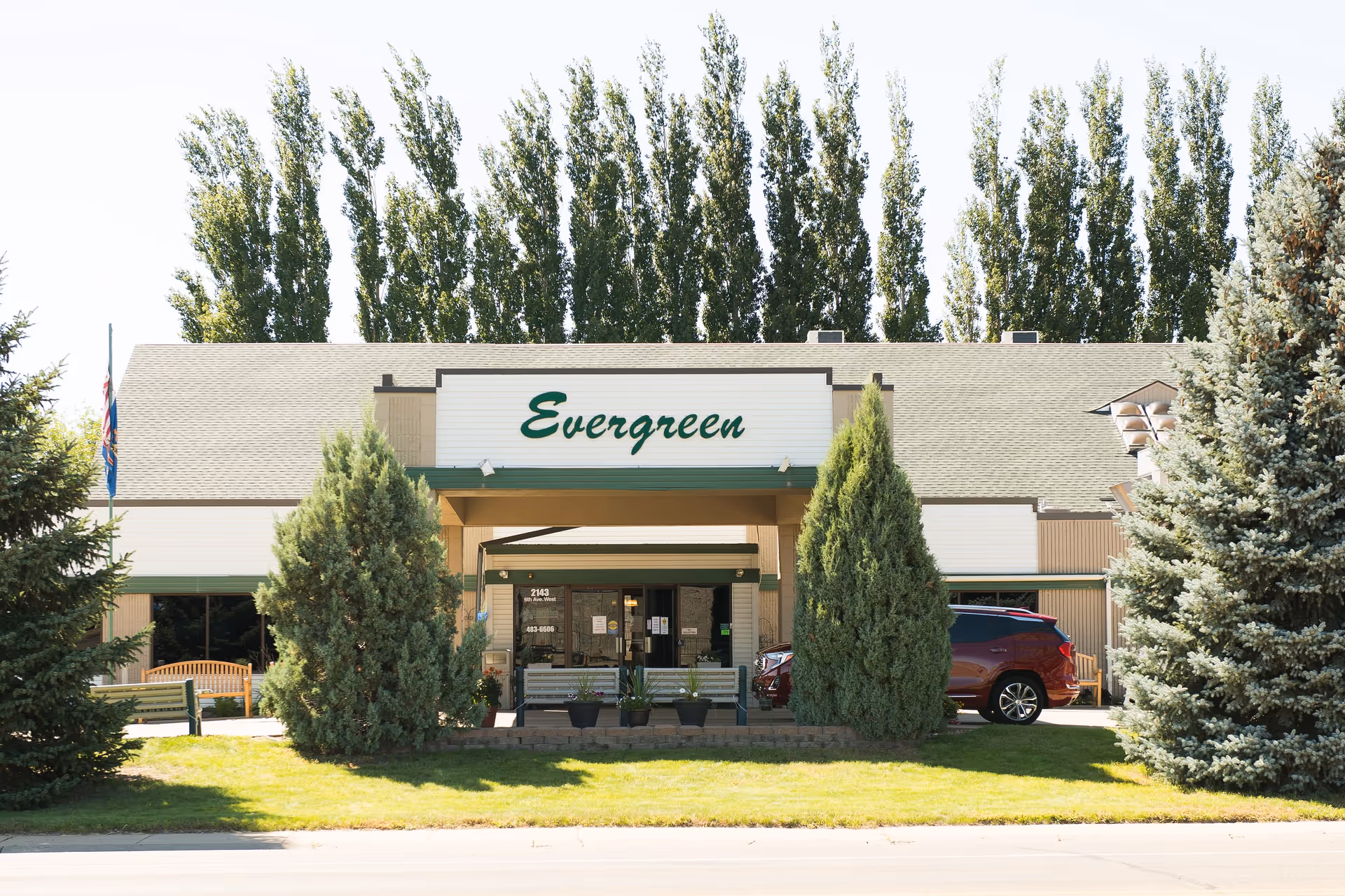 Front exterior view of a single-story building with a sign that reads 'Evergreen' above the entrance. The building is surrounded by tall evergreen trees and has a green lawn in front. There are benches and potted plants near the entrance, and a red vehicle is parked to the right side.