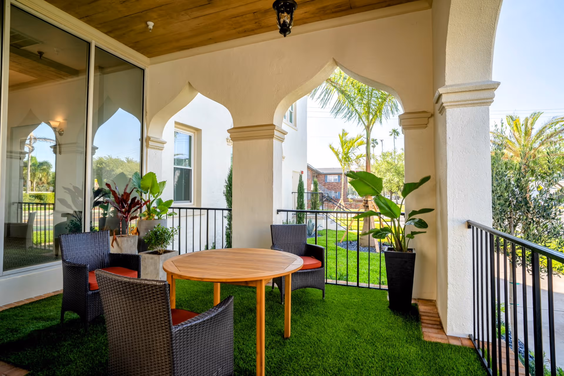 Covered outdoor patio area with a round wooden table and four wicker chairs with red cushions. The patio has artificial grass flooring, potted plants, and arched openings with a view of palm trees and greenery outside.