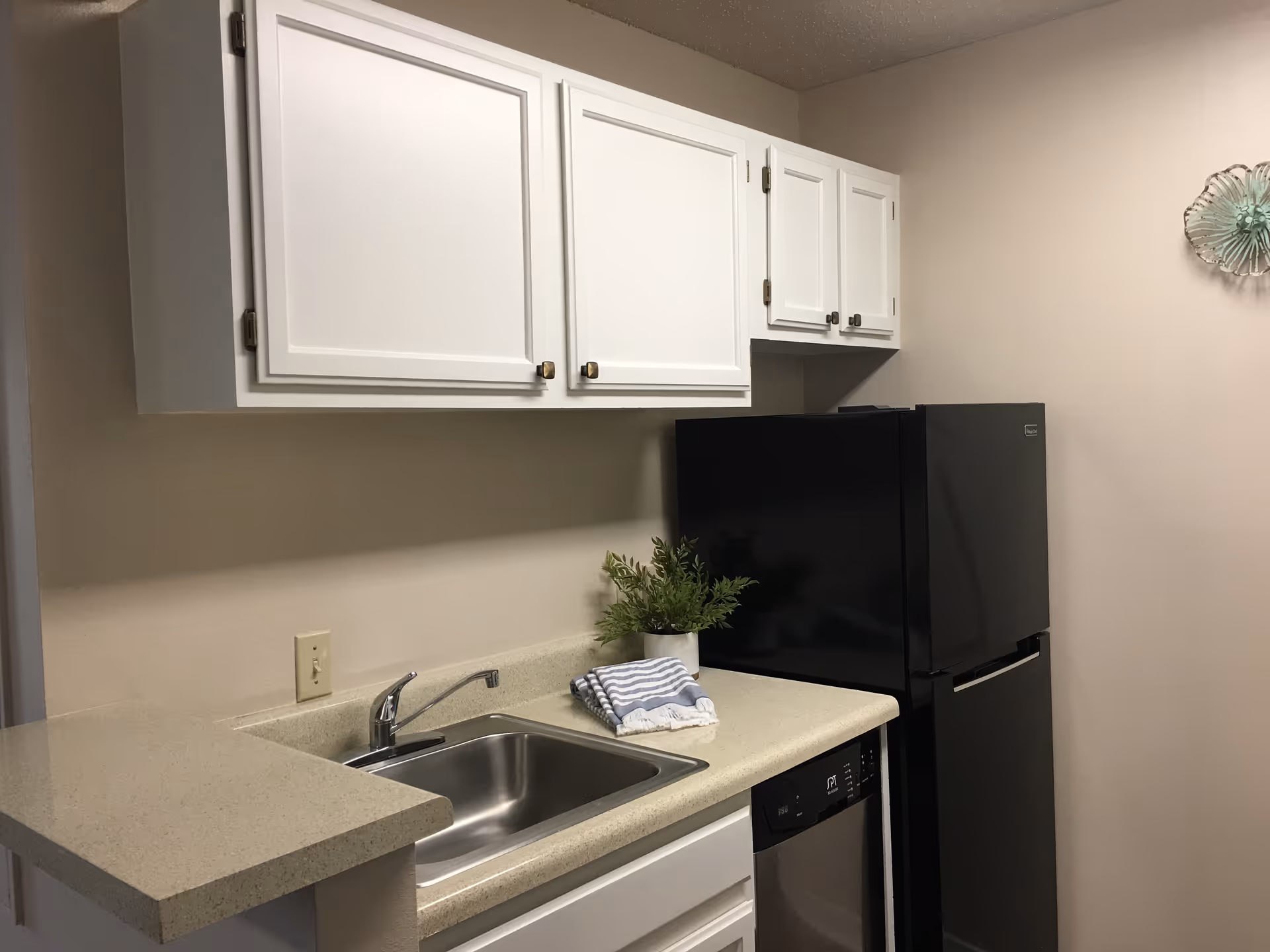 Kitchenette with white upper cabinets, a sink and countertop, a black refrigerator, and a small plant.
