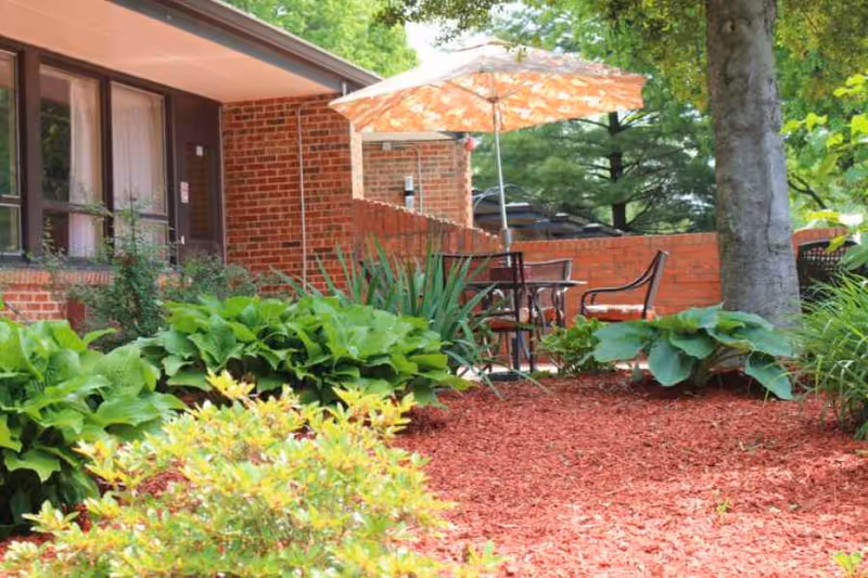 Outdoor patio area with a table, chairs, and an orange patterned umbrella surrounded by green plants and trees next to a brick building.