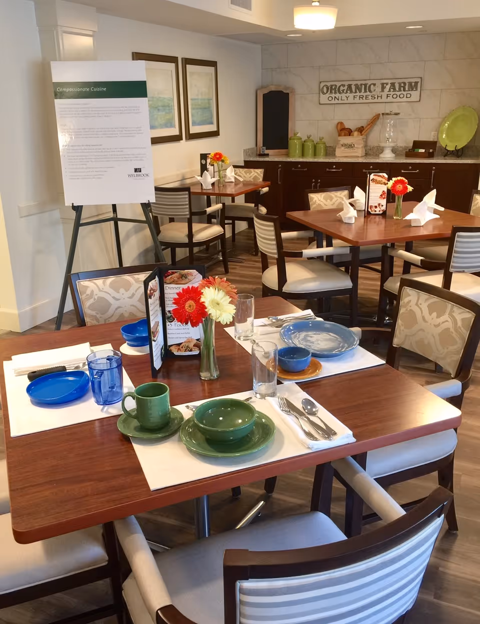 Dining area in a senior living facility with wooden tables set with colorful plates, bowls, cups, and silverware. Each table has a small vase with red and white flowers. In the background, there is a counter with decorative items and a sign that reads 'ORGANIC FARM ONLY FRESH FOOD'. A large informational board is displayed on an easel near the wall.