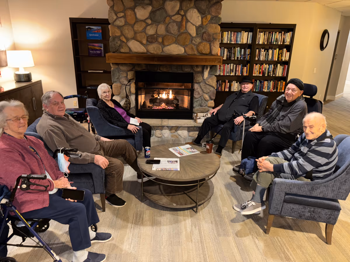 A group of six elderly people sitting in a cozy common area around a round coffee table with a stone fireplace and bookshelves in the background. Some are seated in armchairs and one is in a wheelchair. The room is warmly lit and has a comfortable, inviting atmosphere.