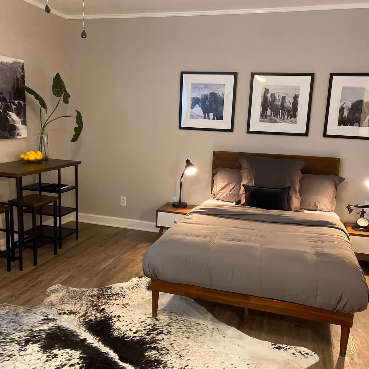 A bedroom with a wooden bed frame and gray bedding, two bedside tables each with a lamp, three black and white framed pictures of horses on the wall above the bed, a small table with two stools on the left side, a vase with large green leaves, and a cowhide rug on the wooden floor.