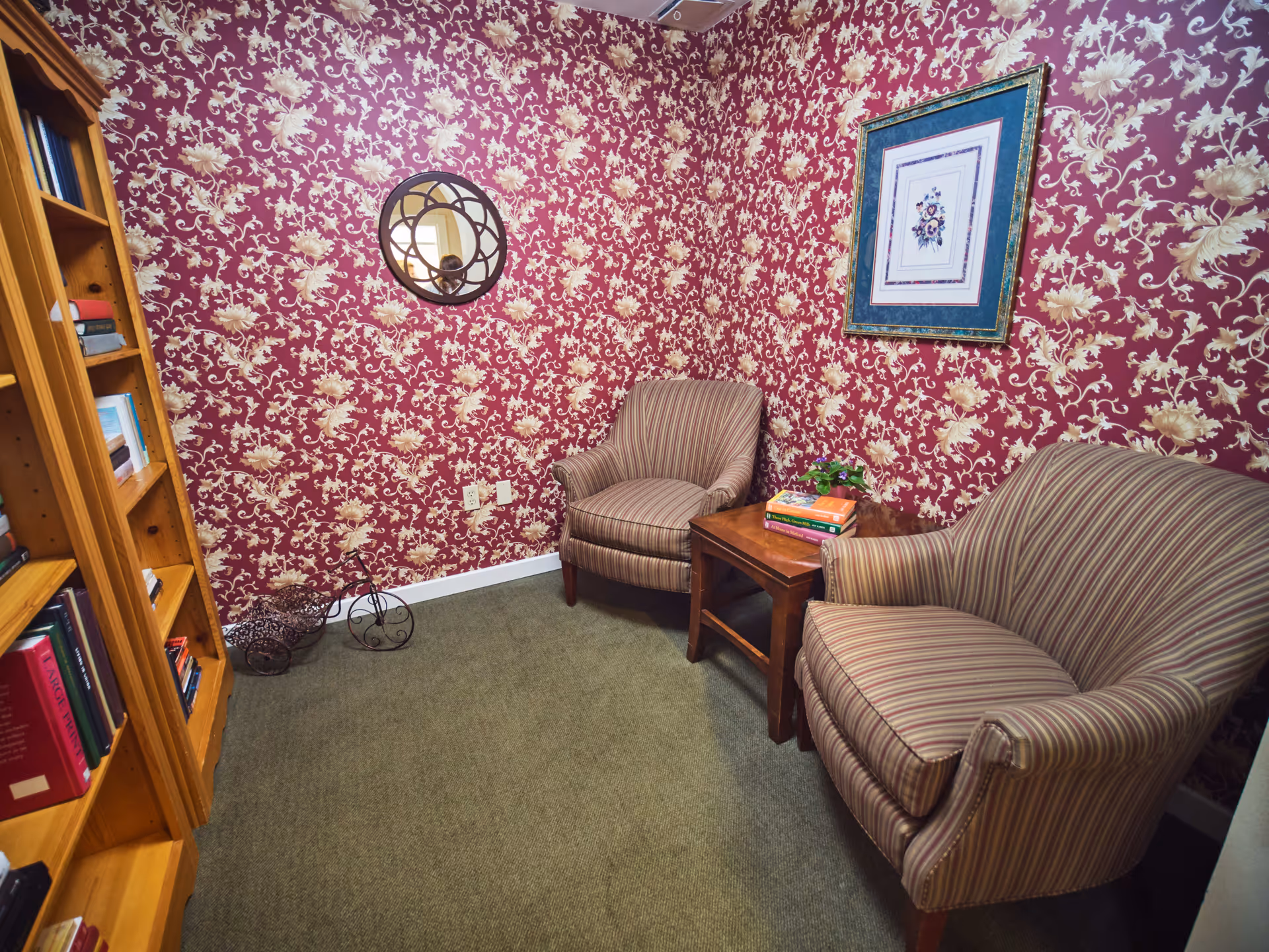 A cozy sitting area with two striped armchairs and a wooden side table with books and a small plant on it. The walls are covered with red floral wallpaper, and there is a round decorative mirror and a framed floral picture hanging on the walls. A wooden bookshelf filled with books is visible on the left side.