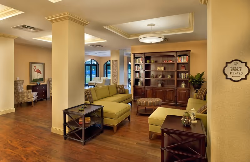 A cozy senior living facility common area with green cushioned sofas, a wooden bookshelf filled with books and decorative items, a round ottoman, and side tables. The room has wooden flooring, beige walls, and a ceiling light fixture. A sign on the wall indicates resident rooms 113-120. In the background, there are additional seating areas with armchairs and large windows letting in natural light.