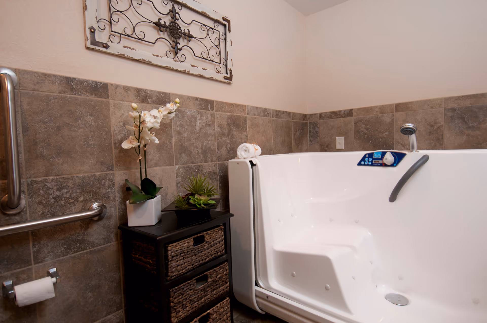 A bathroom with a walk-in bathtub featuring a control panel and a safety handle. The walls are tiled halfway up with brown tiles, and there is a small black cabinet with woven drawers next to the tub. On top of the cabinet are two potted plants, one with white flowers. A decorative metal wall hanging is mounted above the tiles. A grab bar and toilet paper holder are visible on the left side.