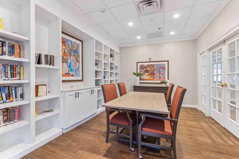 A well-lit room with a wooden table surrounded by six chairs with red cushions. The room features white built-in bookshelves filled with books and decorative items on the left wall, a framed colorful painting above a white cabinet, and a framed landscape photograph on the far wall above a dark wooden sideboard with vases and flowers. The floor is wooden, and there are double glass doors on the right side.
