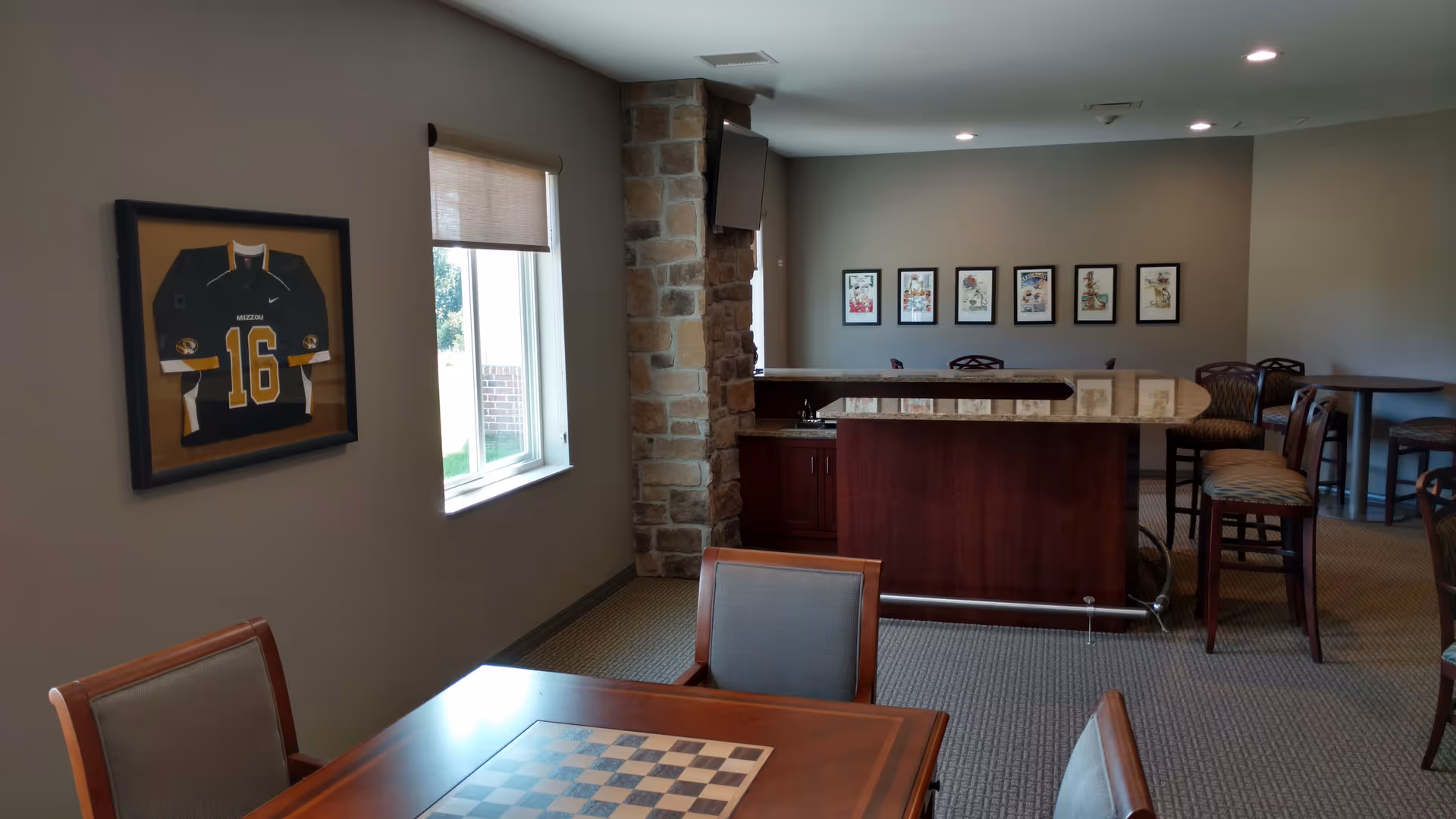 Interior view of a common area in an assisted living facility featuring a wooden table with a built-in checkerboard, several chairs, a small bar with a granite countertop, a mounted TV above a stone pillar, framed pictures on the wall, and a framed sports jersey hanging near a window.
