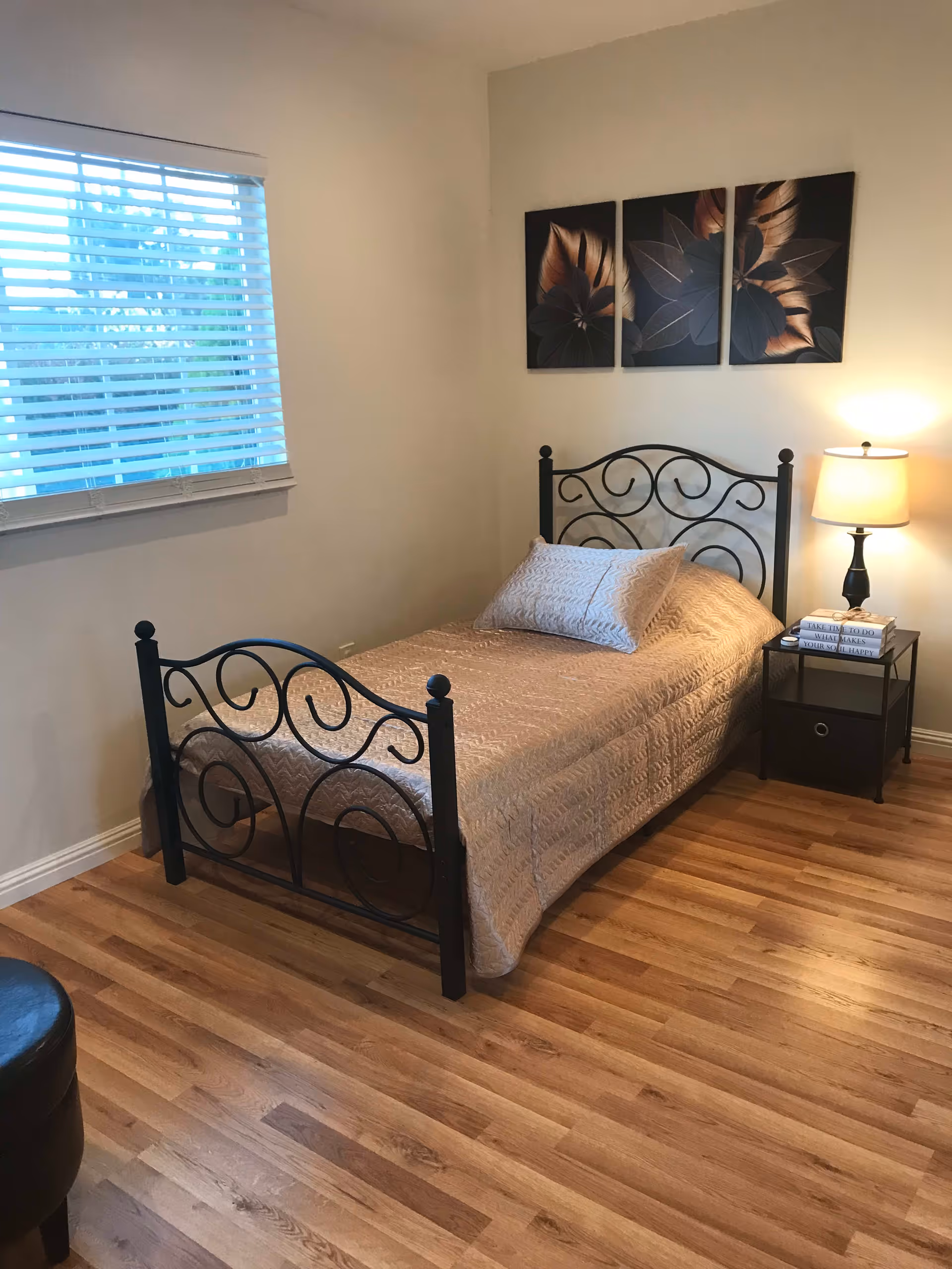 A small bedroom with a single bed featuring a black metal frame and a beige quilted bedspread. There is a pillow on the bed and a nightstand beside it with a lamp and a stack of books. Three decorative floral paintings hang on the wall above the bed. The room has wooden flooring and a window with blinds partially open.