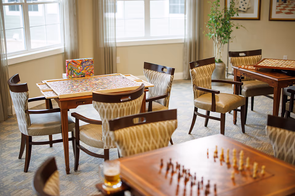 Bright common room with wooden tables and upholstered chairs, a jigsaw puzzle on one table and a chessboard on another by large windows.