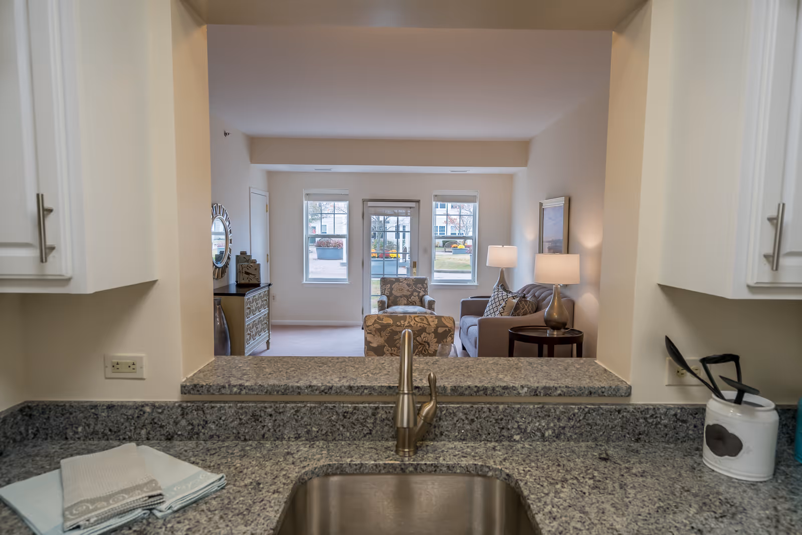 Looking over a kitchen sink and granite countertop through a pass-through into a furnished living room with chairs, lamps, and windows.