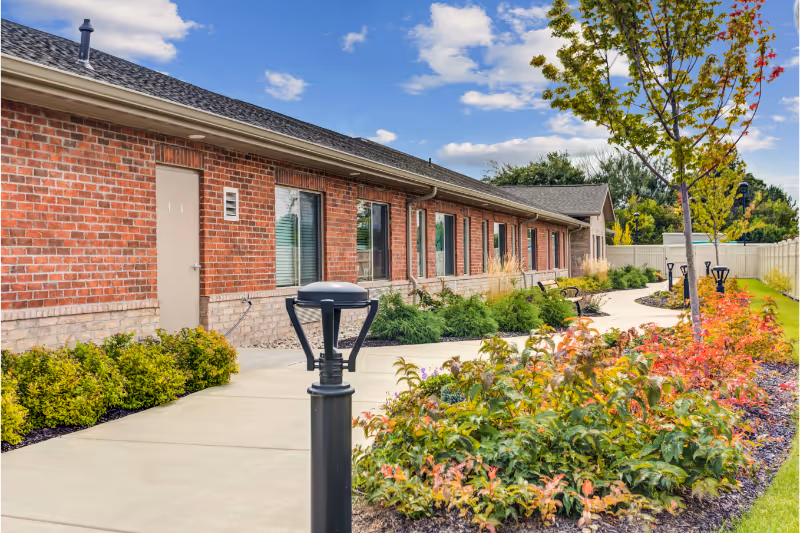 Front exterior of a single-story brick senior living building with a paved walkway, landscaped flower beds, and lamp posts.