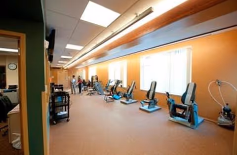 Long interior therapy/exercise room with a row of seated rehabilitation machines along an orange wall and staff in the background.