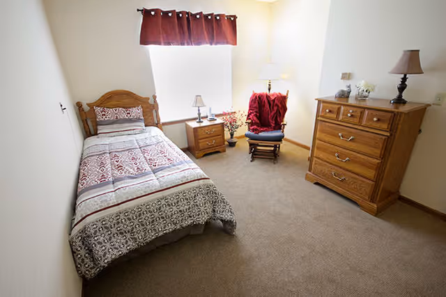 A simple bedroom with a single bed covered in a patterned quilt, a wooden nightstand with a small lamp and decorative items, a wooden rocking chair with a red blanket draped over it, and a wooden dresser with a lamp and flowers on top. The room has beige carpet and white walls with a window covered by a red valance.