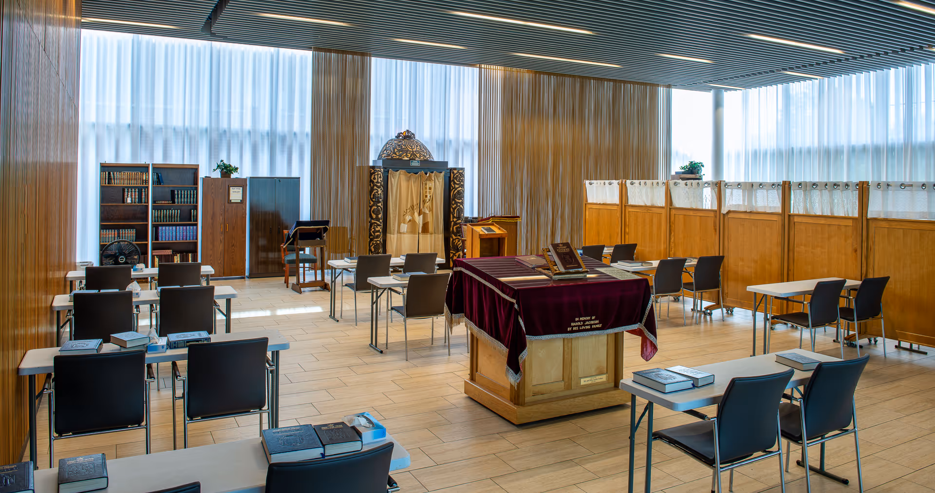 A bright, spacious room with wooden panel walls and large windows covered by sheer white curtains. The room contains several tables and chairs arranged in rows, each table holding books. At the center is a larger table covered with a purple cloth and more books. In the background, there are bookshelves filled with books and a decorative wooden structure with a curtain, suggesting a study or prayer room setting.