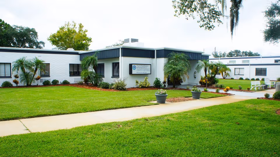 Single-story white nursing facility building with a manicured lawn, palm trees, walkway, and an entrance sign.
