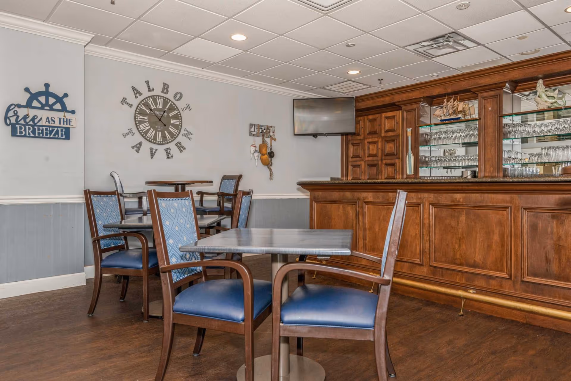 Dining/lounge area with a wooden bar, glassware display, small tables and blue-upholstered chairs, and wall decor including a large clock.