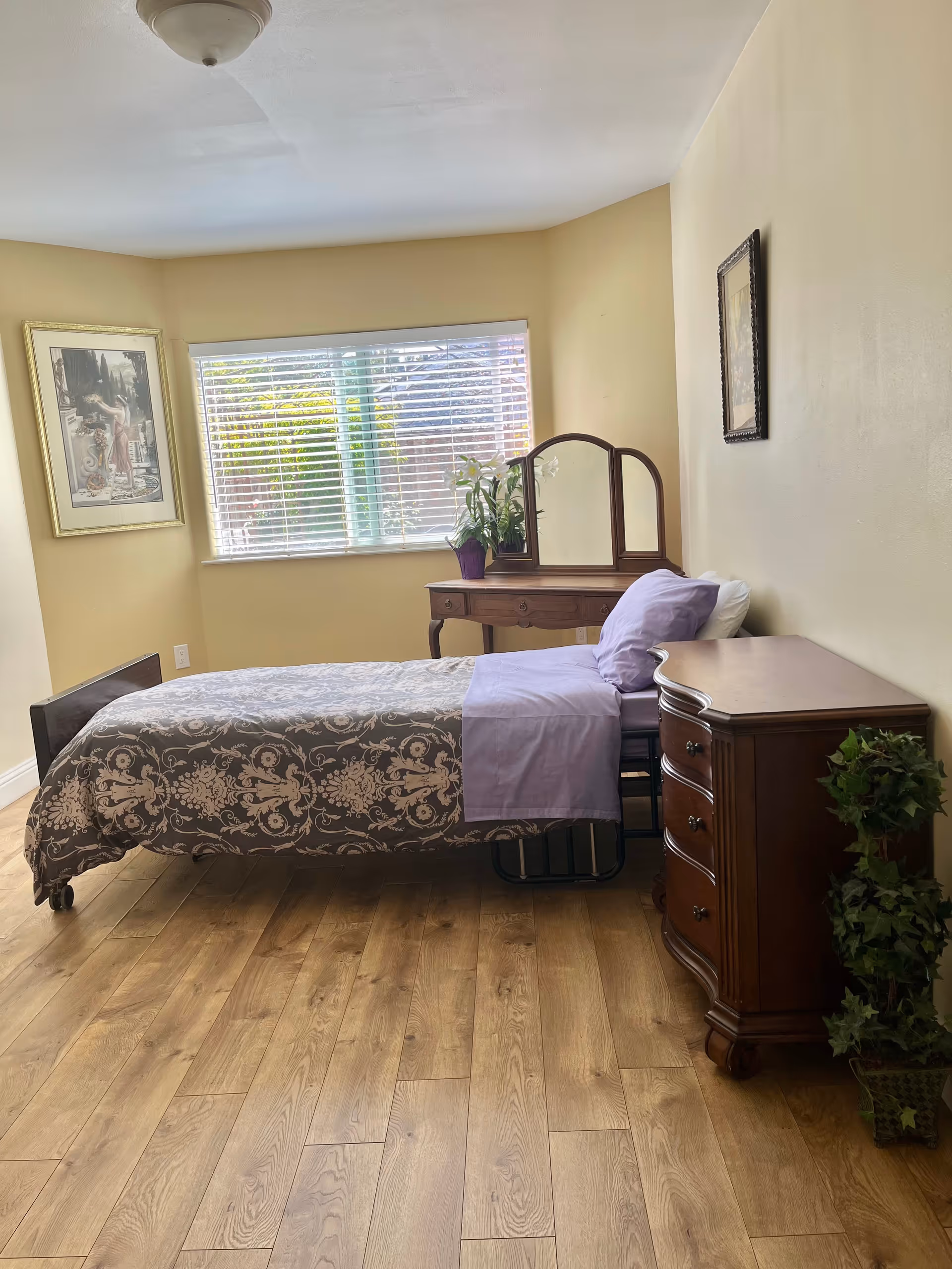A bedroom with a single bed covered in a patterned gray and white bedspread and a lavender sheet and pillow. There is a wooden dresser with a potted plant next to it, and a wooden vanity with a mirror and a potted plant on top. A window with white blinds lets in natural light, and framed artwork hangs on the walls. The floor is wooden.