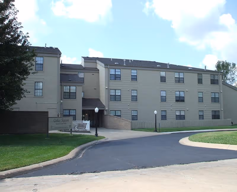 Exterior view of a three-story beige building with multiple windows, a curved driveway, green lawn, and a sign that reads Cedar Square near the entrance.