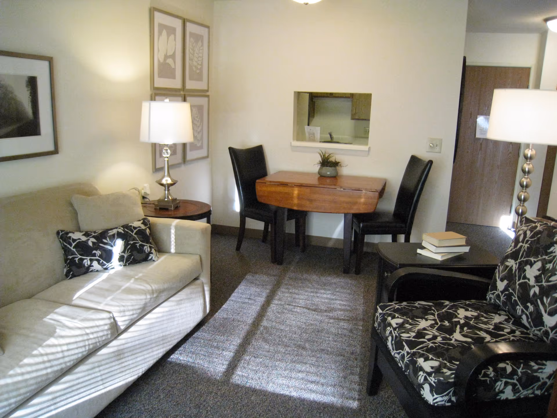 Cozy living room with a sofa, patterned armchair, floor lamps, and a small dining table by a kitchen pass-through.