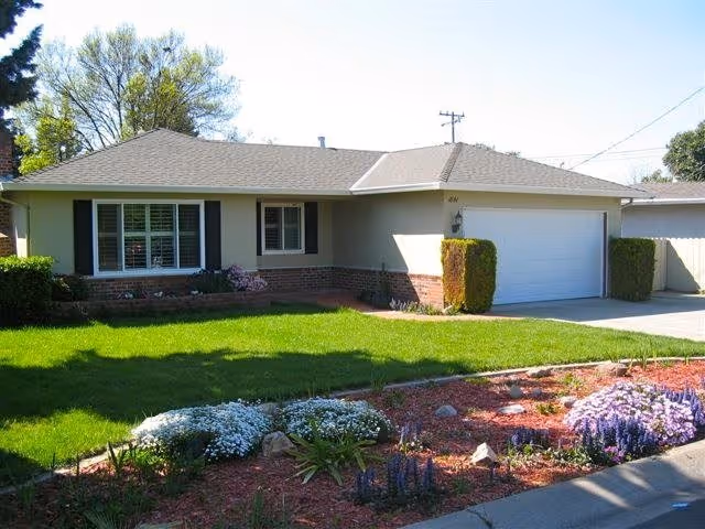 Single-story house with a well-maintained front lawn and garden featuring green grass, flowering plants, and shrubs. The house has a light-colored exterior with a two-car garage and a large window with shutters.