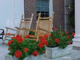 Two wooden rocking chairs sit on a front porch beside red geranium planters.
