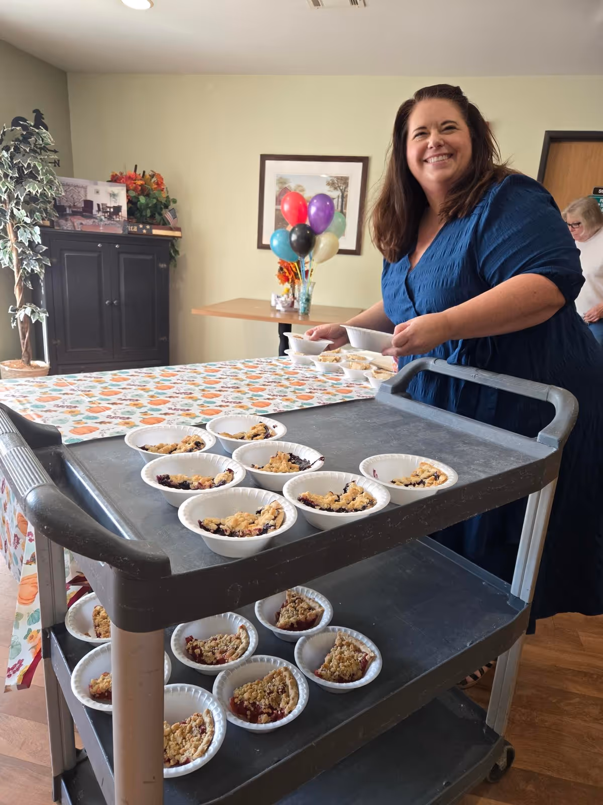 A smiling woman in a blue dress stands behind a serving cart filled with small bowls of dessert in a room decorated with a colorful tablecloth, balloons, and a potted plant. Another person is partially visible in the background near a door.