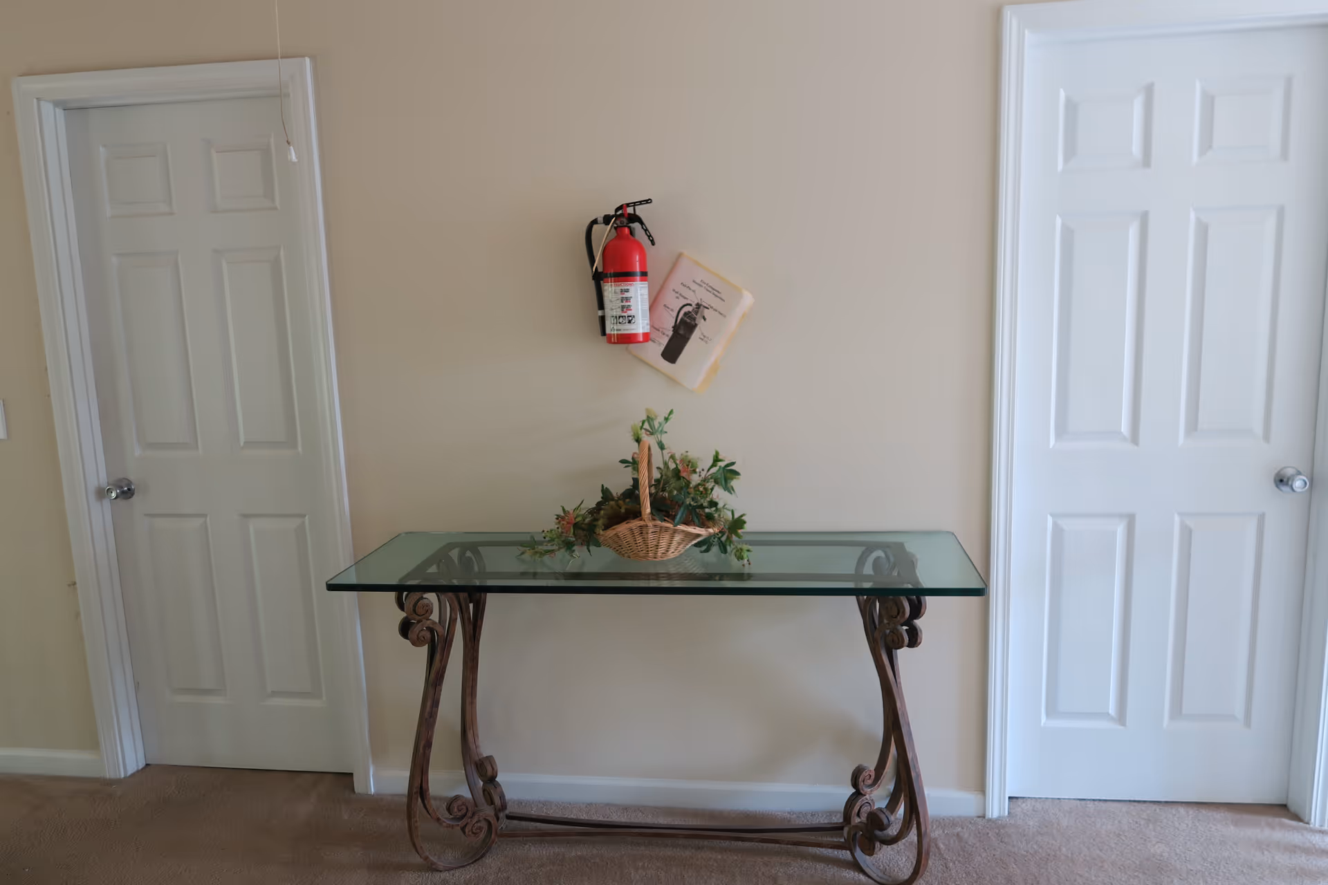 A glass-top table with ornate metal legs is placed against a beige wall between two white paneled doors. On the table is a wicker basket filled with green and pink artificial flowers. Above the table on the wall is a red fire extinguisher with an instruction sheet attached behind it.