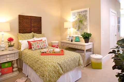 A cozy bedroom with a single bed featuring a green patterned bedspread and multiple decorative pillows. There is a wooden headboard, two bedside tables with lamps and decorative items, a tray with a teapot and cups on the bed, a potted plant on a dresser, and light-colored walls with framed artwork.