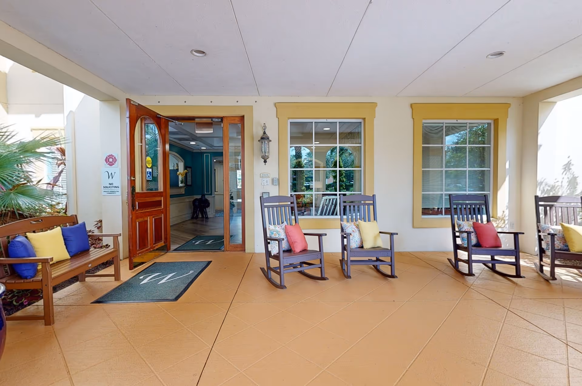 Covered outdoor seating area at the entrance of The Windsor of Lakewood Ranch featuring wooden rocking chairs with colorful cushions and a wooden bench with pillows. The entrance door is open, showing a glimpse of the interior with teal walls and decorative elements.