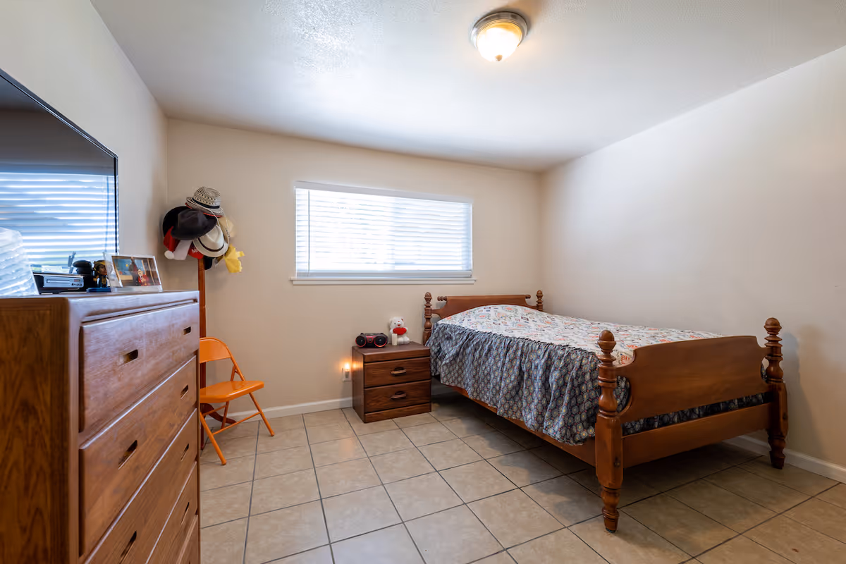 A simple bedroom with a wooden bed covered with a patterned bedspread, a wooden nightstand with a radio and a stuffed toy, a wooden dresser with framed photos and a TV on top, an orange folding chair, and a coat rack holding several hats. The room has tiled flooring, beige walls, and a window with blinds letting in natural light.