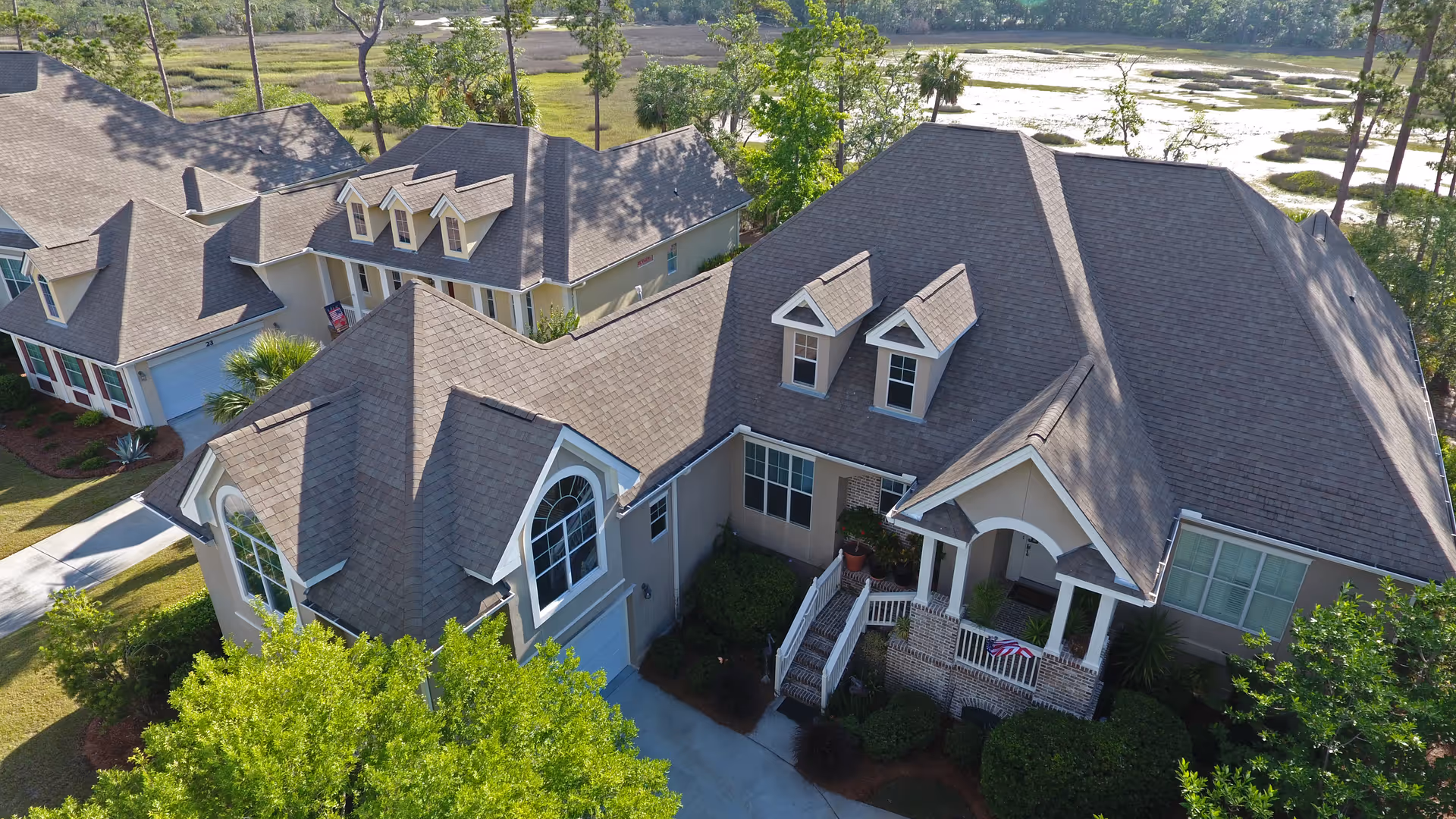 Aerial view of a residential-style building with a large porch, multiple dormer windows, and a well-maintained garden. The building is surrounded by greenery and situated near a marshland area.
